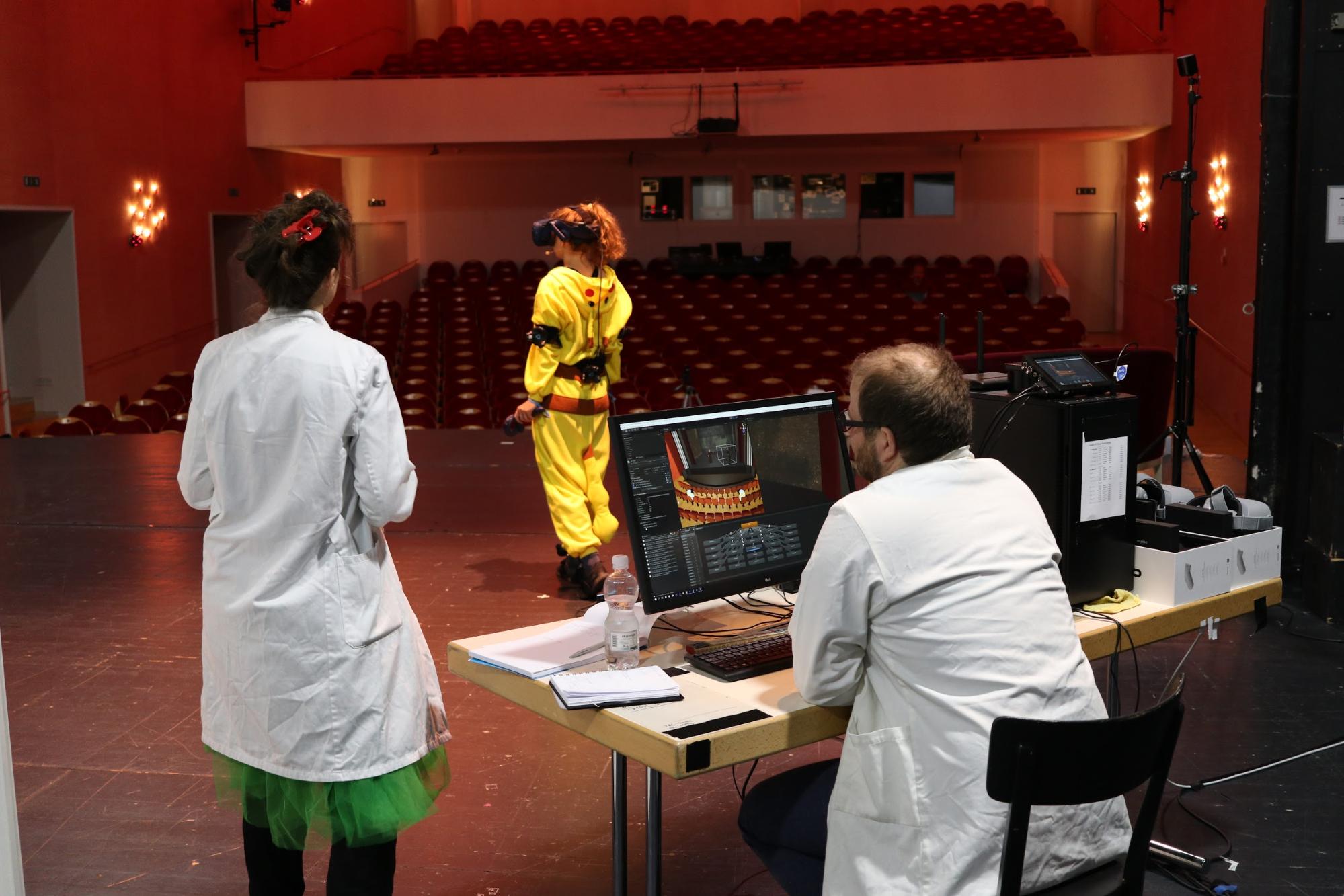 Woman in yellow using VR while others watch her and a screen