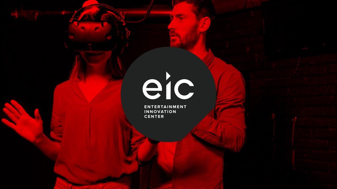 Person with a virtual reality headset standing next to another person in a dark room with a red hue overlaid on the whole image with the logo and words, "Entertainment Innovation Center".