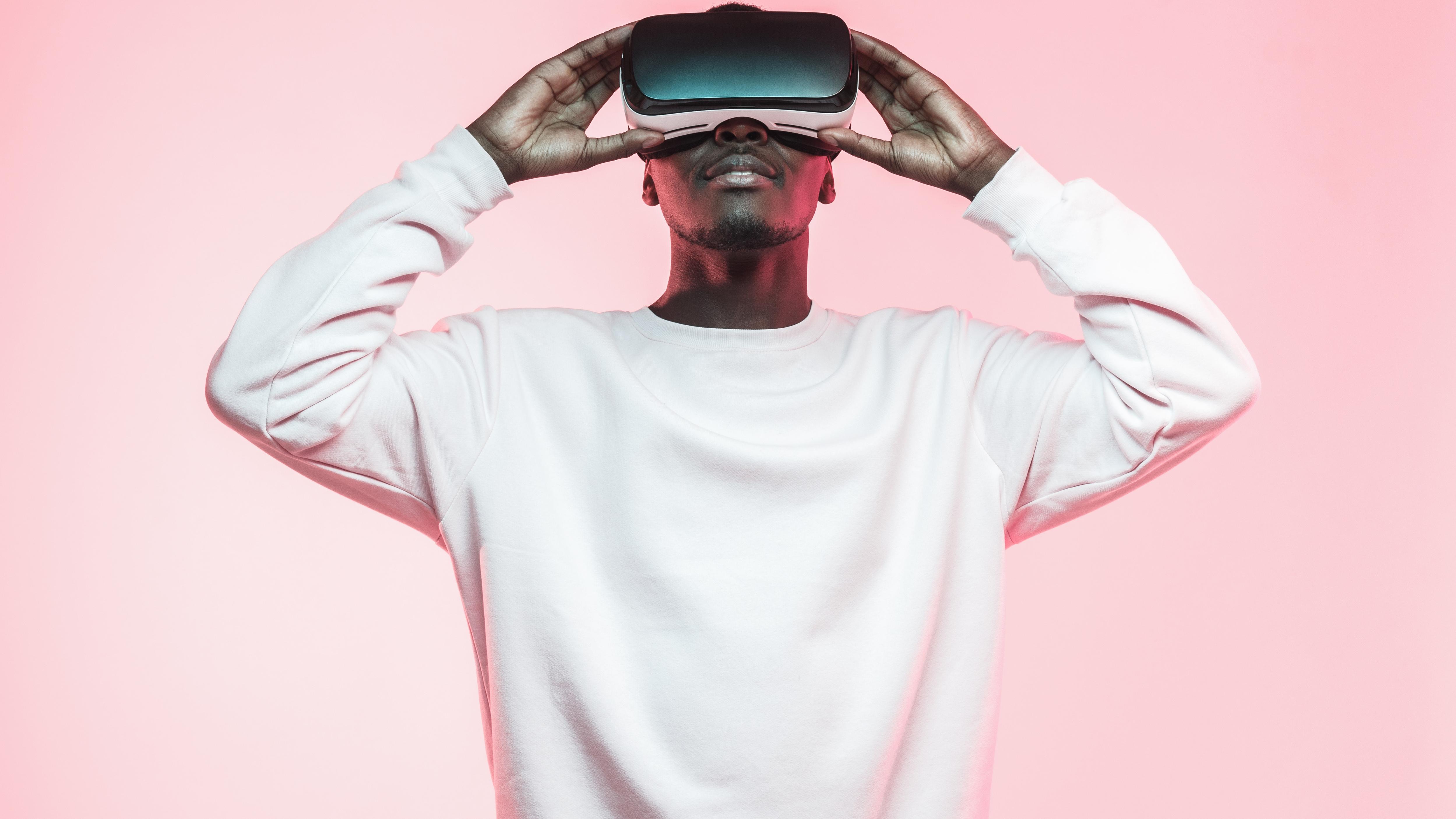 Young african man holding virtual reality goggles with both hands, isolated on pink background