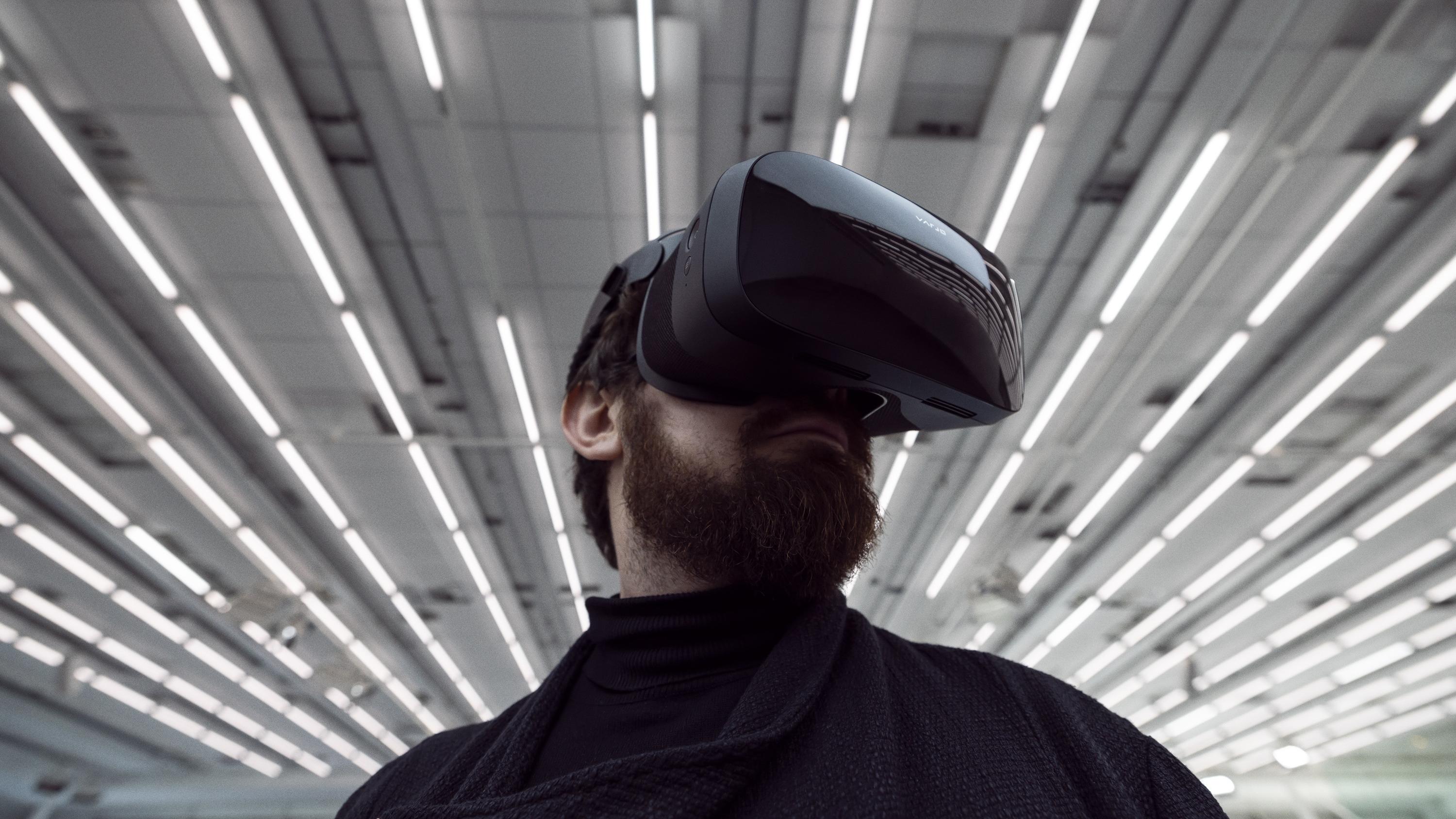Person wearing a mixed reality headset. The background is industrial tube lighting on the ceiling. The image shows the person from the shoulders up.