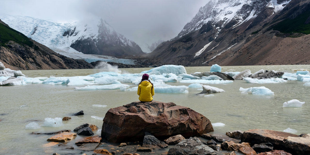 In the foreground there is a person with long brown hair and a yellow coat sitting on a large rock surrounded be smaller rocks. In the background there is chunks of ice melting into a body of water and mountains tipped with snow.