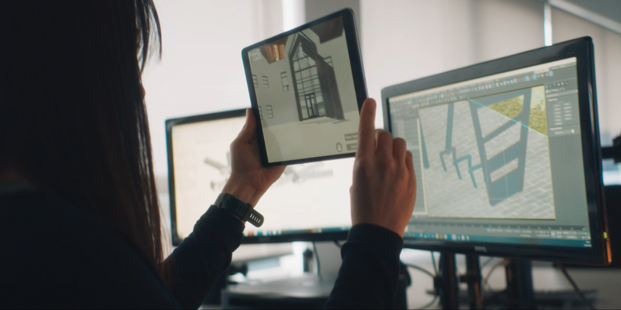 Person holding up tablet showing plans of a building. They are sitting in front of two PC screens. The on the left has an something on it that is difficult to see and the one on the right has the Unity editor open and in the editor is the building. 