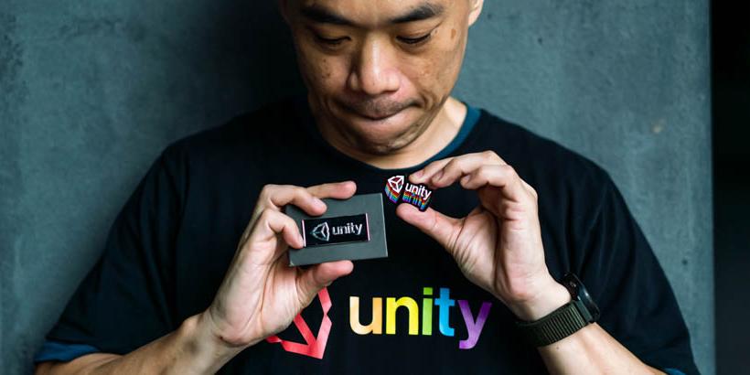 A person in a black Unity t-shirt with rainbow-colored text looks down at two Unity badges they are holding.