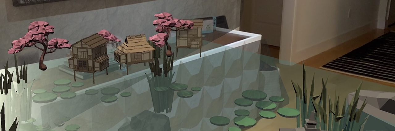 An animated pond with lily pads, rocks surrounding an in it and tall grass dotted around. One one side their are little wooden Japanese-style houses and pink cherry blossom trees. On the other there is a wooden Japanese-style tower. This animated scene has been created in AR and is being displayed on the ground and part of the wall of a room with light wooden floors and white walls.