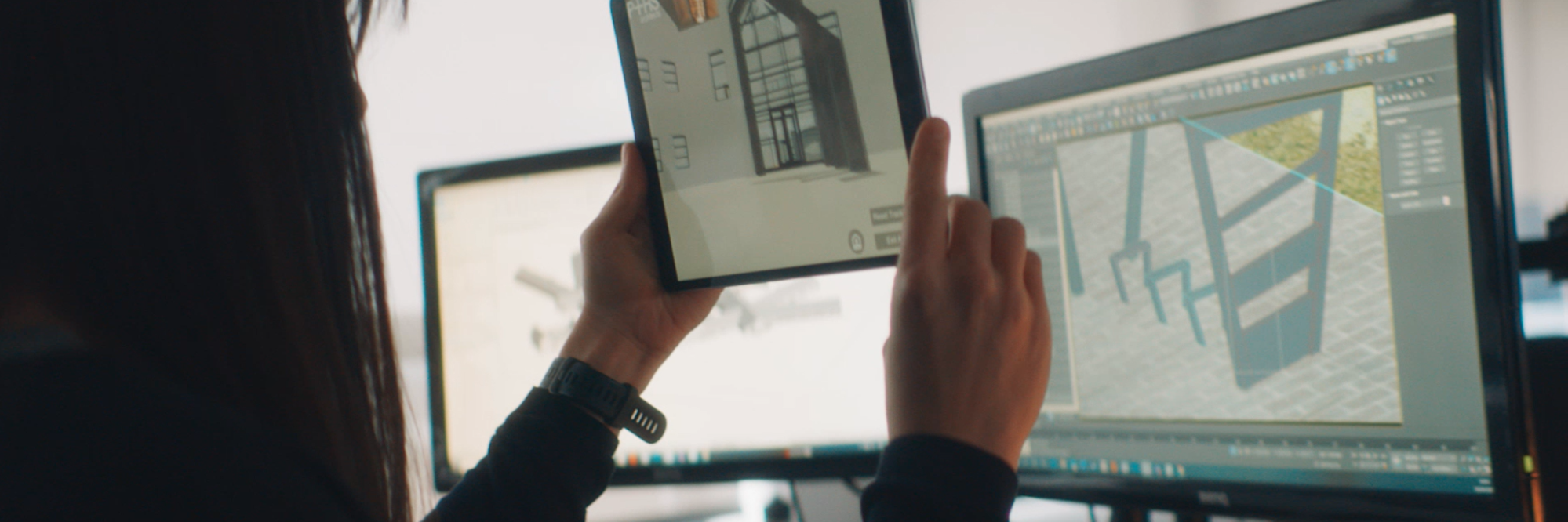 Person holding up tablet showing plans of a building. They are sitting in front of two PC screens. The on the left has an something on it that is difficult to see and the one on the right has the Unity editor open and in the editor is the building.