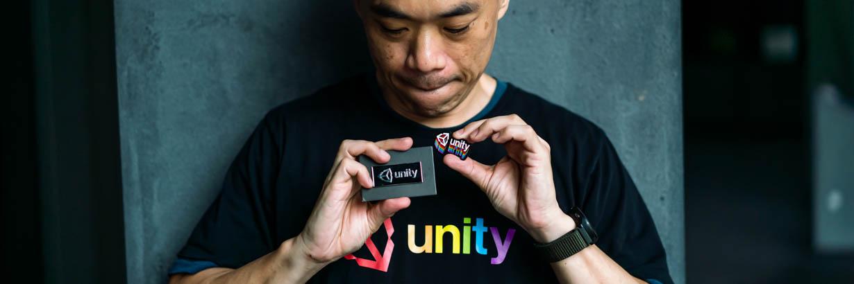 A person in a black Unity t-shirt with rainbow-colored text looks down at two Unity badges they are holding.